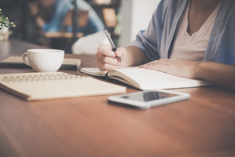 A woman writes Article in a notebook at a café table with a coffee and smartphone nearby.